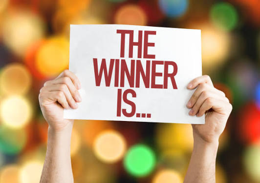 A pair of hands hold up a sign that reads "The winner is" in front of a twinkling colorful backdrop